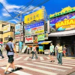 Scorcio urbano di Okinawa in Yakuza Kiwami 3 con pedoni che attraversano la strada e insegne colorate.