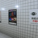 Corridoio di The Exit 8 con poster sul muro e cartello della telecamera di sicurezza con occhi stilizzati.