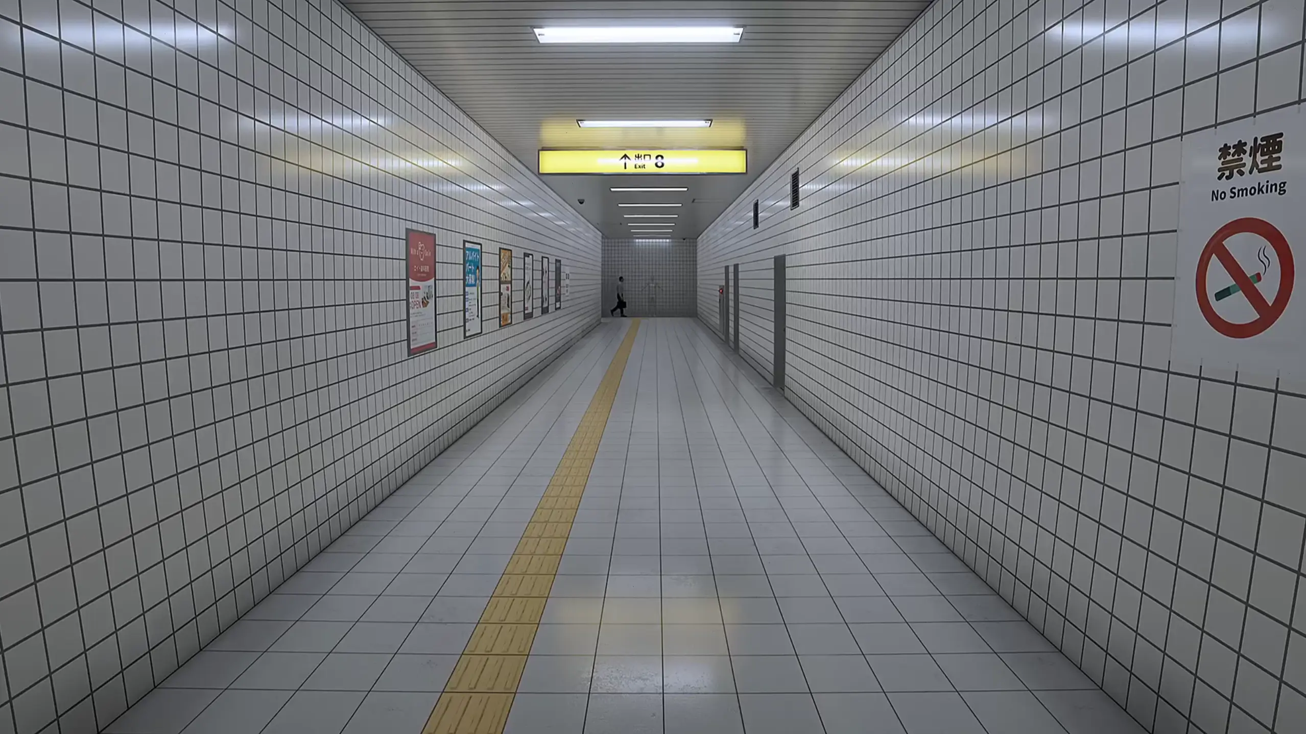 Lungo corridoio piastrellato con poster alle pareti e un cartello giallo di direzione in fondo.