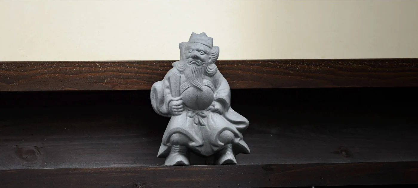 Piccola statua grigia in pietra di Shōki, la versione giapponese del Cacciatore di Demoni, posizionata su una trave di legno scuro.